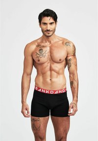 Svarta kalsonger med ett rött "FRANK DANDY"-midjeband, åtsittande design, slät textur och en modell som visar upp olika tatuerade överkroppar.