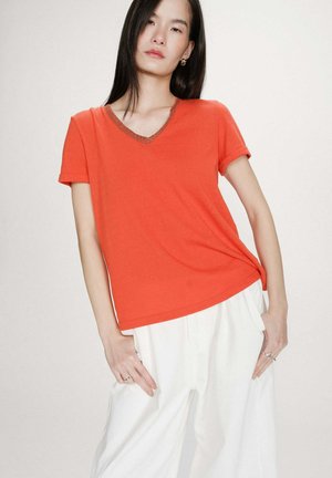 Femme aux longs cheveux bruns portant un t-shirt orange à manches courtes et col en V, ainsi qu'un pantalon blanc à jambes larges, debout devant un fond clair uni.