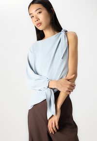 Blusa di seta azzurro chiaro con un design asimmetrico, dettaglio nodo su una spalla e manica lunga e fluida con polsino annodato. Abbinata a pantaloni marroni.