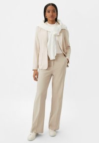 Blazer beige sur-mesure avec poche, pantalon beige clair assorti, pull en maille blanc drapé et baskets blanches. Textures de tissu lisses et élégantes.