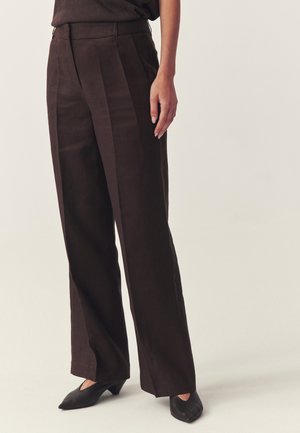 Kobieta ubrana w ciemnobrązowe szerokie spodnie z wysokim stanem i czarne buty z szpiczastym noskiem, stojąca na tle jednolitego jasnego tła.