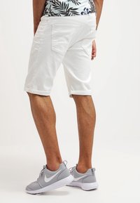 Shorts en jean blanc avec des ourlets retroussés, dotés de poches arrière et d'une texture lisse. Portés avec des chaussures de sport grises.