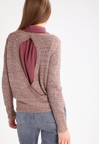 Hellbrauner Strickpullover mit strukturierter Oberfläche, mit einem großen offenen Rücken, getragen über einem bordeauxroten Oberteil, kombiniert mit grauen Jeans.