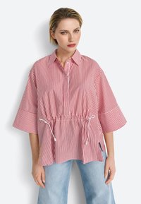 Maglietta a righe rosse e bianche con colletto, maniche corte e vita regolabile con cordoncino. Tessuto morbido, vestibilità comoda e spacchi laterali.