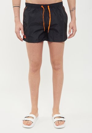 Mannelijke benen met zwarte zwemshorts met oranje trekkoorden en logo, en witte sandalen met dubbele bandjes, staand op een witte achtergrond.