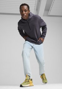 Sudadera gris de manga larga con capucha y cremallera, jeans azul claro y zapatillas deportivas verde-negro con acentos amarillos.