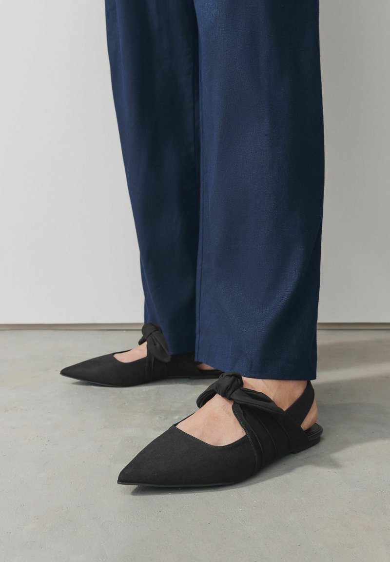 Pantofi negri din suede, cu toc slingback și vârf ascuțit, având un detaliu cu curea legată și un design cu profil scăzut. Purtați cu pantaloni bleumarin.