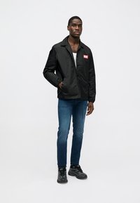 Μαύρο μπουφάν με κολάρο και κόκκινο λογότυπο, φορεμένο πάνω από άσπρο πουκάμισο, σε συνδυασμό με μπλε τζιν και μαύρα παπούτσια αθλητικά.