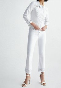 Chaqueta de mezclilla blanca con detalles de ojales en los hombros y puños, combinada con jeans blancos acampanados y zapatos de tacón blancos con tiras.