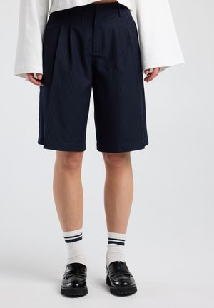 Persoon draagt marineblauwe knielange pinstripe short, witte sokken met zwarte strepen, zwarte loafers en een witte top met lange mouwen.