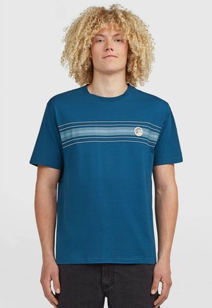 Young person with curly blonde hair wearing a blue t-shirt with horizontal stripes and a circular logo on the chest, standing against a plain background.