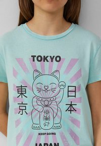 Lichtblauw katoenen T-shirt met een afbeelding van een glimlachende kat, Japanse karakters en een zonnestraalpatroon in lavendel en roze.