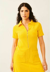 Keltainen lyhythihainen mekko, jossa on vetoketjukiinnitys edessä, kaulus ja kaksi päällyspocketsia. Valmistettu pehmeästä kankaasta, jolla on sileä pinta.