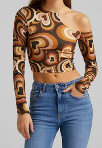 Brown patterned crop top with long sleeves, featuring a one-shoulder design and abstract heart shapes in earthy tones. Paired with blue jeans.