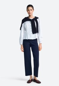 Camisa blanca abotonada con cintura fruncida, pantalones azul marino y suéter de punto oscuro sobre los hombros. Zapatos tejidos color burdeos. Texturas suaves.