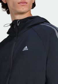 Čierna bunda na zips s kapucňou, s reflexným logom Adidas na hrudi a kontrastnými pruhmi na ramenách. Hladká textúra.