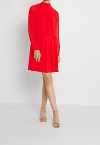 Punavuoren pitkäpunainen mekko, jossa on korkea kaula-aukko, levenevä helma ja läpikuultavat yksityiskohdat hartioilla. Yhdistetty kultaisiin hihna-sandaaleihin.