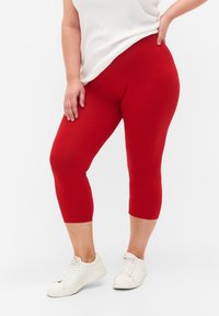 Červené legíny capri vyrobené z elastického materiálu, s hladkou texturou, těsným střihem a elastickým pasem. Nosené s bílými teniskami.