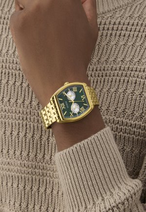 Gold metal watch with a green face, Roman numerals, and two sub-dials, set on a wrist covered by a knitted beige sweater.