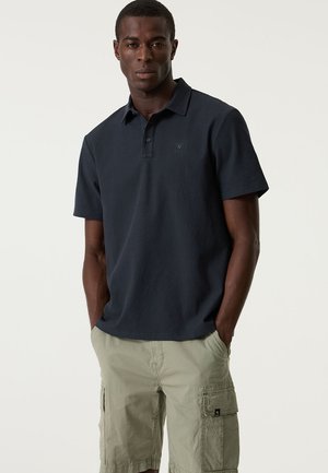 Polo blu navy con design strutturato, maniche corte e abbottonatura con tre bottoni, abbinata a pantaloncini cargo verde chiaro.