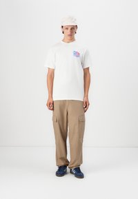 White cotton t-shirt with colorful graphic print on left chest, paired with tan cargo trousers featuring pocket details, and blue sneakers.