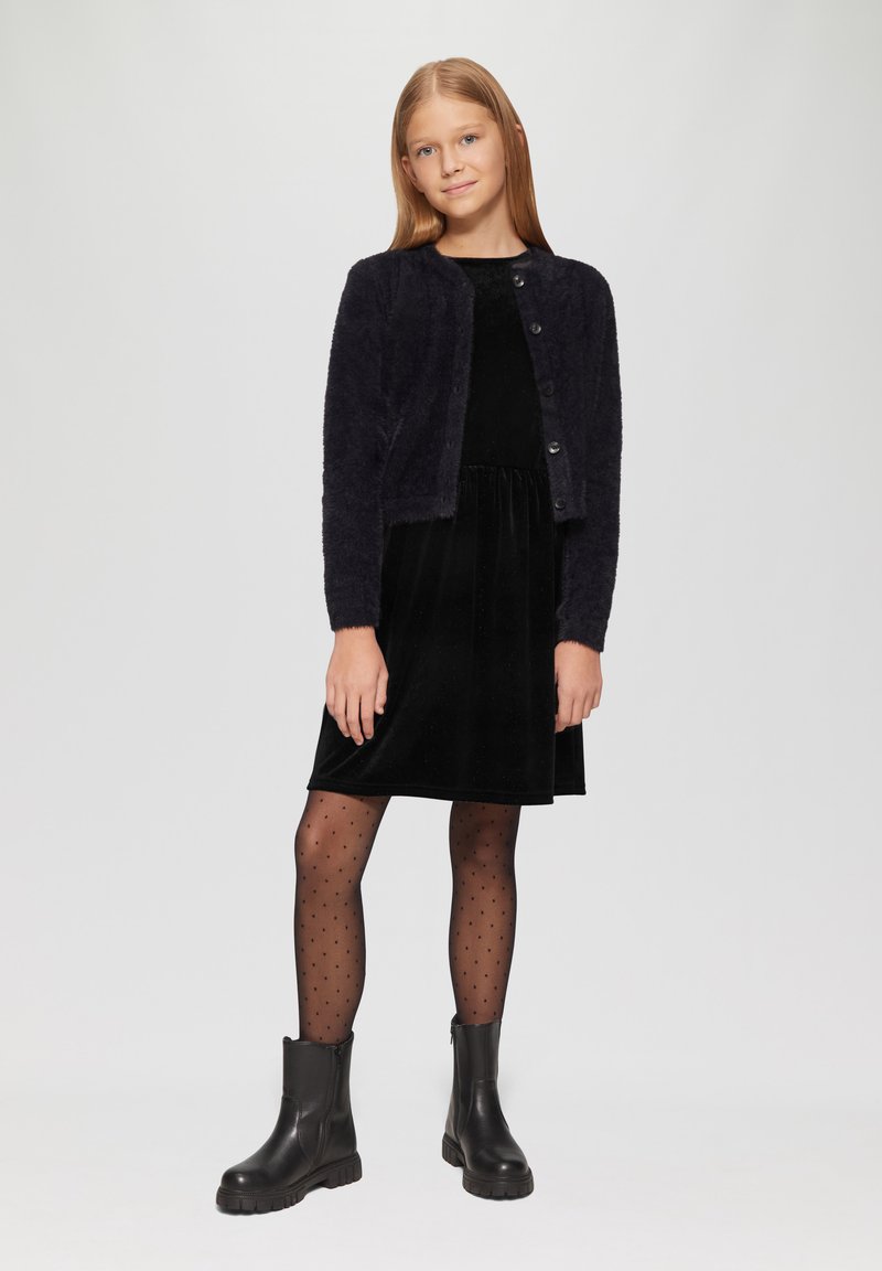 Marineblauer strukturierter Cardigan mit Rundhalsausschnitt und Knopfverschluss, kombiniert mit einem schwarzen Samtkleid und gepunkteten Strumpfhosen, abgerundet mit schwarzen Stiefeletten.