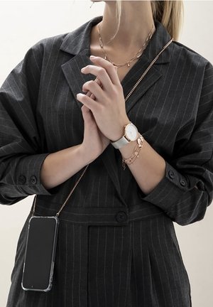 Blazer noir à fines rayures avec manches retroussées, montre argentée et bijoux avec accents roses. Smartphone dans une coque transparente suspendu à une chaîne.
