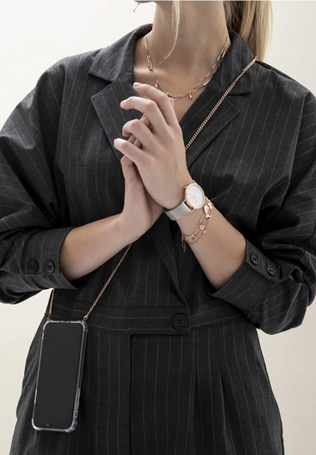 Blazer noir à fines rayures avec manches retroussées, montre argentée et bijoux avec accents roses. Smartphone dans une coque transparente suspendu à une chaîne.