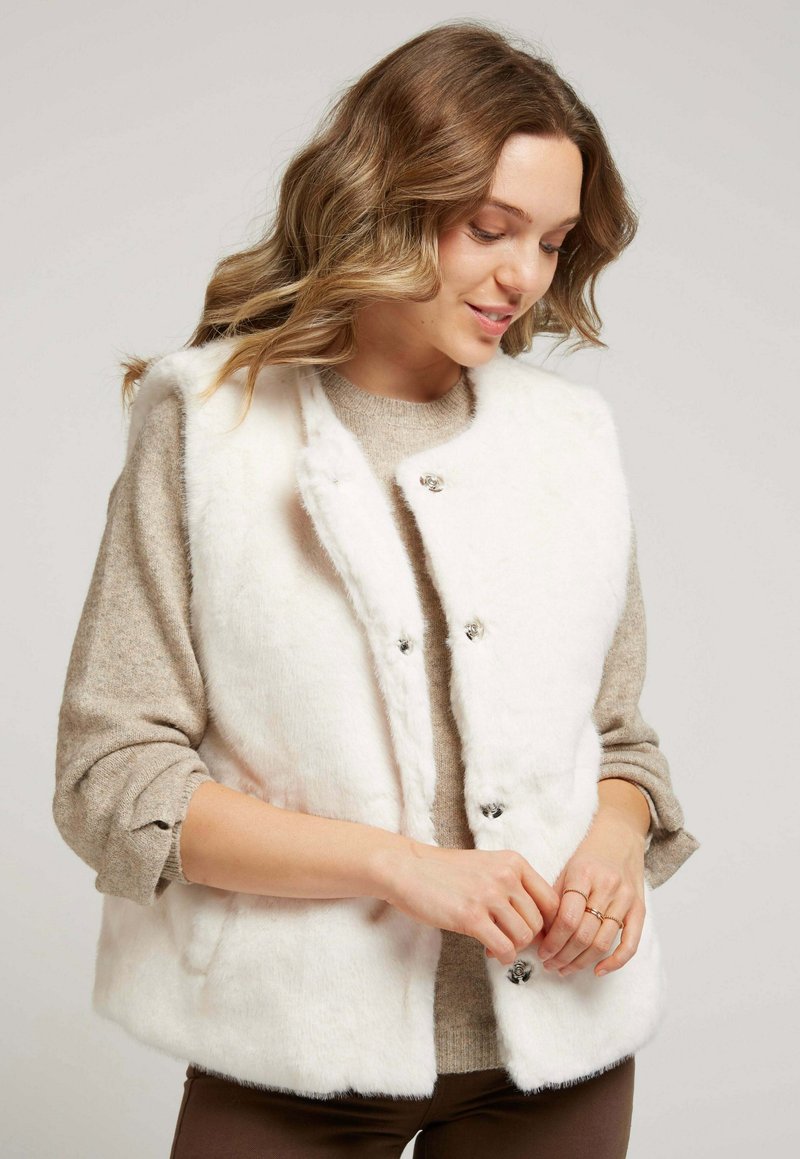 Gilet en fausse fourrure blanche avec une texture douce, col rond et fermetures à pression argentées. Porté sur un pull marron clair, mettant en valeur le jeu de couches.