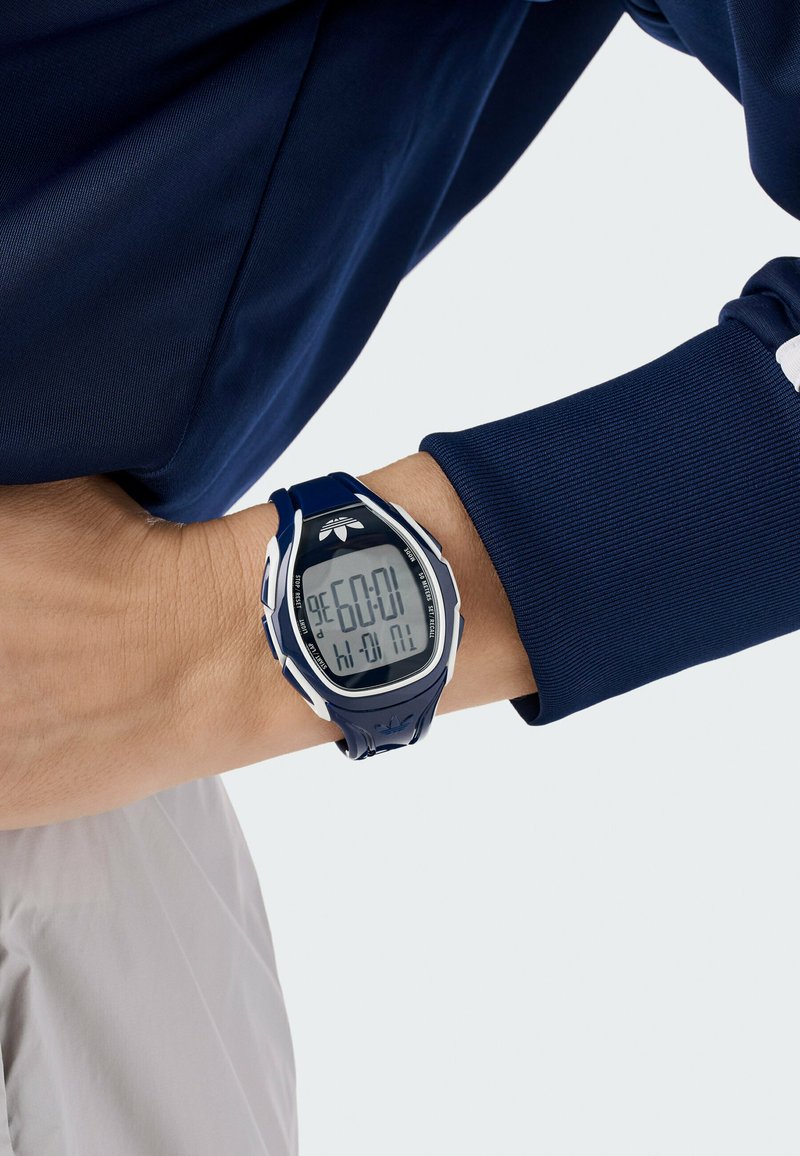 Orologio digitale sportivo con cassa e cinturino in plastica blu navy. Lo schermo LCD mostra l'ora e presenta accenti bianchi. Indossato su un polso con una camicia blu navy.