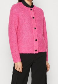 Plyschig rosa cardigan med långa ärmar, rund hals och fem svarta knappar framtill. Mjuk struktur och något oversize-passform.