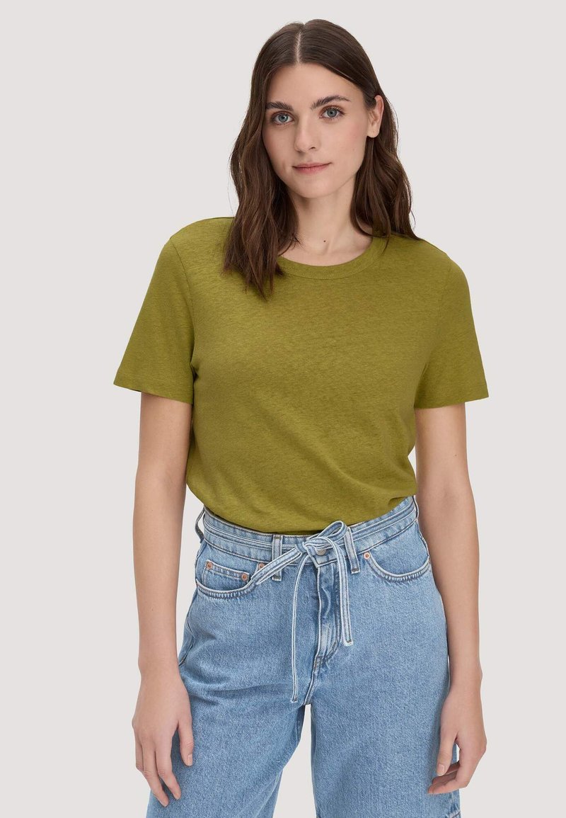 Femme aux cheveux bruns mi-longs portant un t-shirt kaki à manches courtes rentré dans un jean bleu clair taille haute avec une ceinture en tissu.