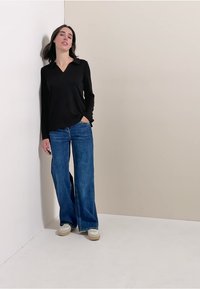 Czarna bluzka z długim rękawem i kołnierzykiem, w połączeniu z rozszerzanymi niebieskimi dżinsami. Stylizacja cechuje luźny krój i minimalne wykończenia. Widać casualowe obuwie.