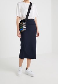 Marineblauwe kokerrok tot de knie van rekbaar materiaal, gecombineerd met een los witte top en een gestreepte crossbody tas. Witte platform sneakers.