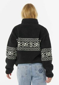 Giacca in pile nera con dettagli a motivo bianco su petto e maniche, colletto alto e vestibilità comoda, abbinata a jeans blu chiaro.