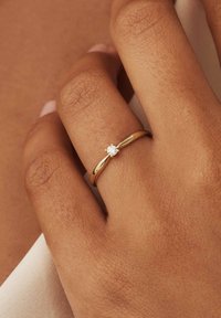 Gouden ring met een centrale ronde diamant, een gladde band en subtiele groeven voor textuur. Hand toont een natuurlijke huidskleur en detail.