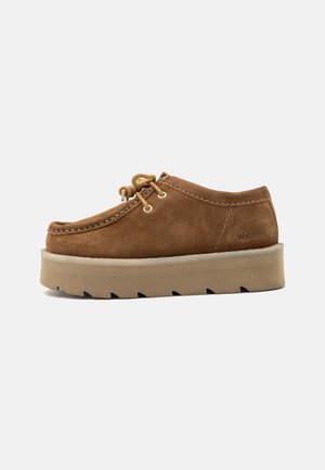 Chaussure décontractée en daim marron avec semelle épaisse couleur fauve, lacets noués à l'avant et logo "Wallabees" sur le côté.