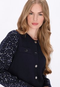 Tengeri pulóver, flitterekkel díszített ujjal, gombos elülső résszel és zsebfedéllel a mellkason. Simább textúrával és fényes díszítésekkel.