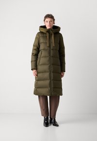 Persona vestida con un abrigo acolchado largo de color verde oliva con capucha, pantalones marrones y botas negras hasta el tobillo, de pie contra un fondo blanco liso.