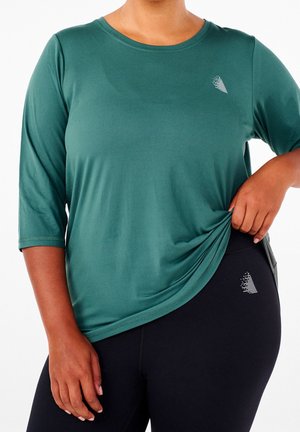 Osoba ubrana w zielony sportowy top z długim rękawem i czarne legginsy, trzymająca bok topu, oba ubrania mają małe białe logo.