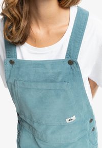 Tyrkysové manšestrové overaly se širokými ramínky, přední kapsou a černými knoflíky. Nositeli přes bílý tričko. Obsahuje logovou nášivku.
