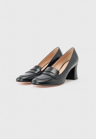 Mocassins pour femmes en cuir noir avec talons carrés, bouts en forme d'amande et design sans lacets sur un fond clair.
