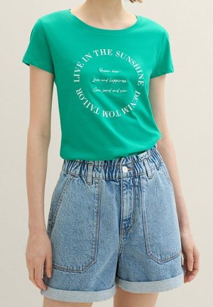 Mujer con camiseta verde con texto blanco circular y shorts vaqueros de tiro alto y color azul claro con los bajos remangados.