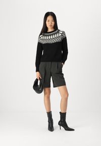 Schwarzer Pullover mit weißem geometrischen Muster, graue maßgeschneiderte Shorts, schwarze Knöchelstiefel, hält eine kleine schwarze Handtasche, auf einem neutralen Hintergrund.