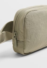 Borsa a tracolla color khaki chiaro con forma rettangolare, tessuto a trama liscia, chiusura con zip e tracolla in tessuto. Dettagli di cucitura lungo i bordi.
