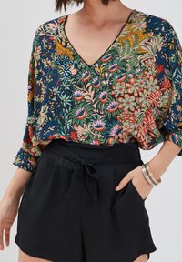 Koszulka z krótkim rękawem w kwiatowy wzór w wyrazistych kolorach, z dekoltem w kształcie litery V i luźnym krojem, zestawiona z czarnymi szortami o wysokim stanie. Widoczne srebrne bransoletki.