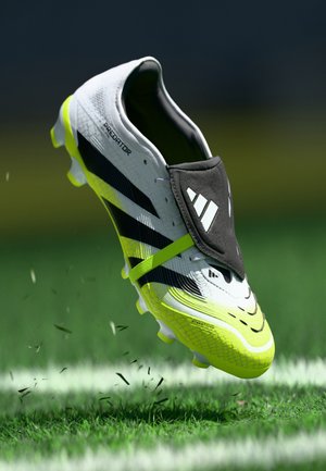 White and neon yellow Adidas Predator soccer cleat kicking up grass on a green field with a black lace cover partially lifted.