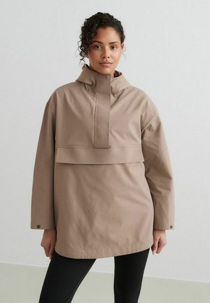 Vrouw draagt een oversized beige jas met kap en voorflap en zwarte legging, staand tegen een effen lichte achtergrond.