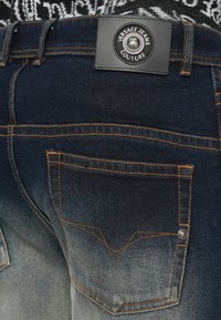 Tamno plave oprane traperice s narančastim šavovima, detaljima na stražnjim džepovima i crnom oznakom Versace Jeans Couture na pojasu.