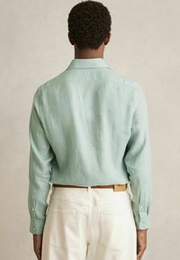 Cămașă din in verde deschis cu mâneci lungi, cu manșete cu butoni și o croială lejeră. Asortată cu pantaloni albi prevăzuți cu buzunare pe spate.